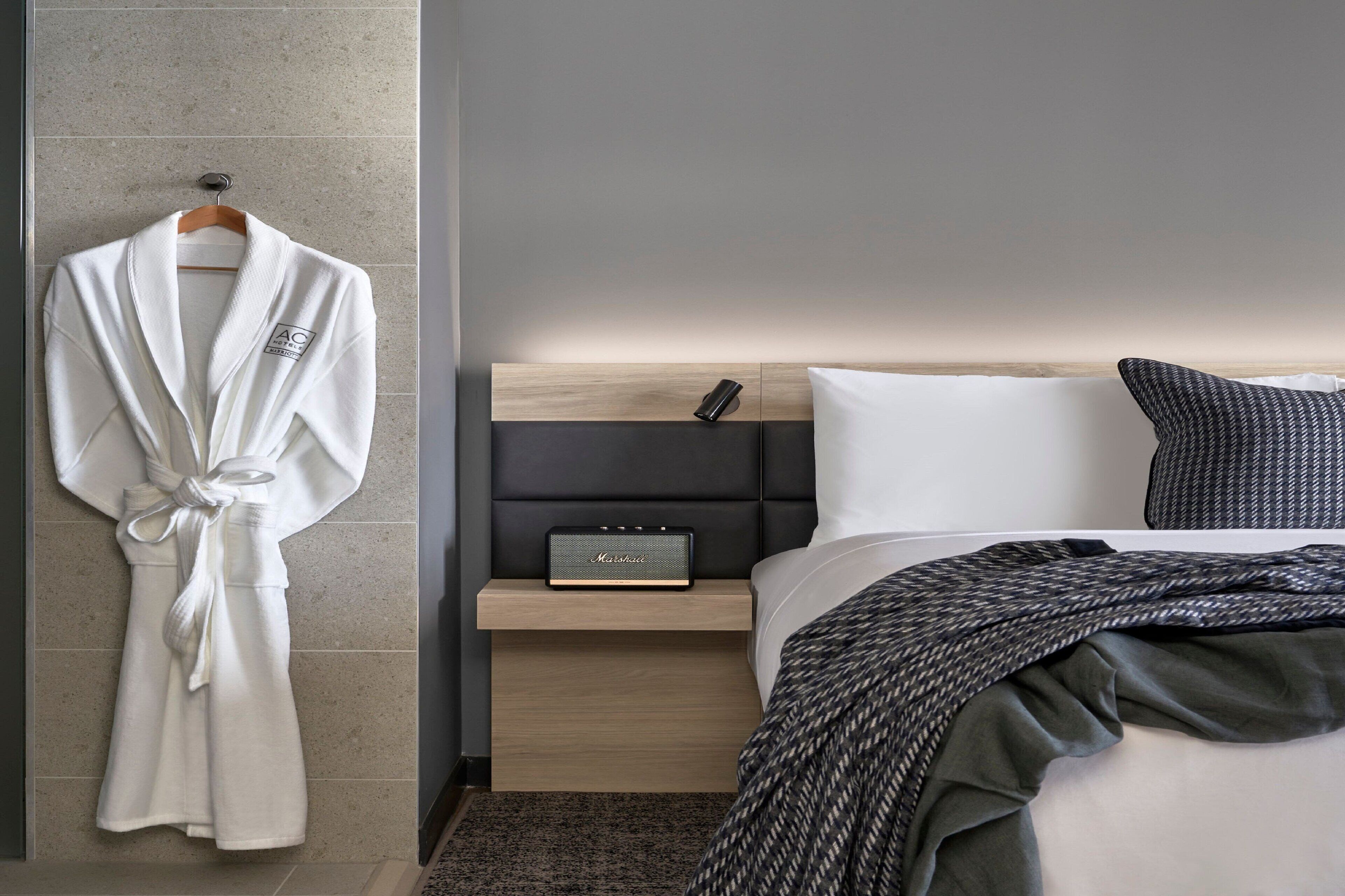 A modern hotel room with a neatly made bed, a bedside table with a radio, and a robe hanging on the wall.