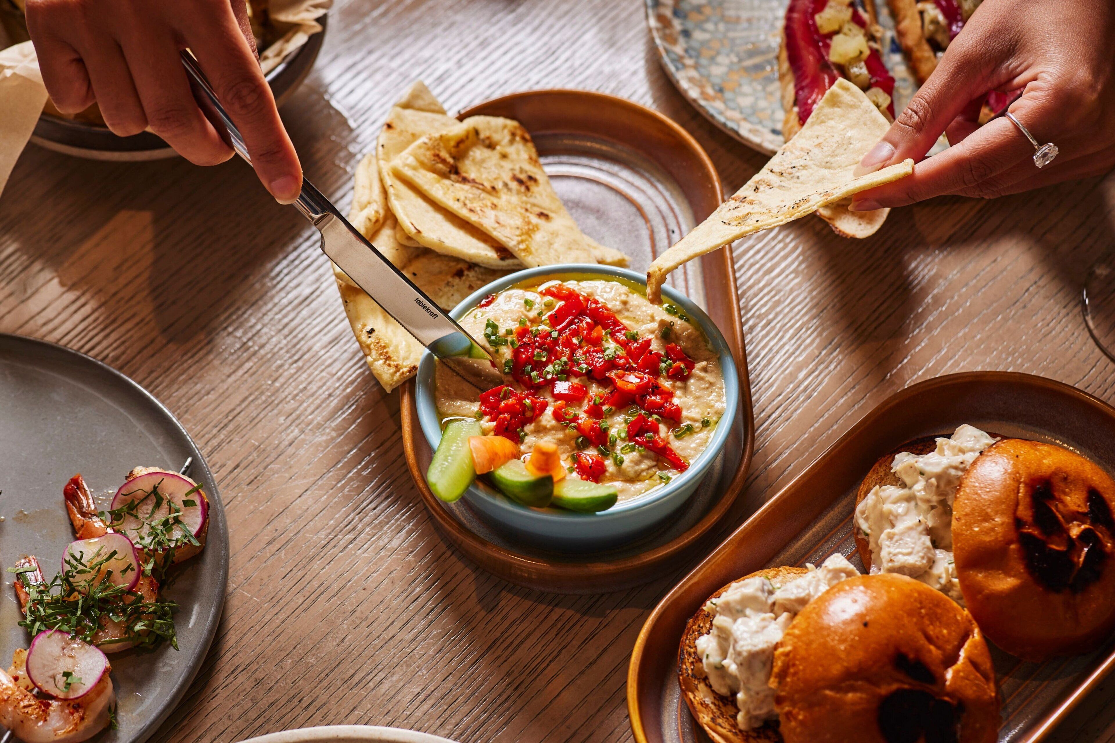 Een eettafel met diverse gerechten, waaronder een kom hummus versierd met groenten, platbrood en een bord gamba's.