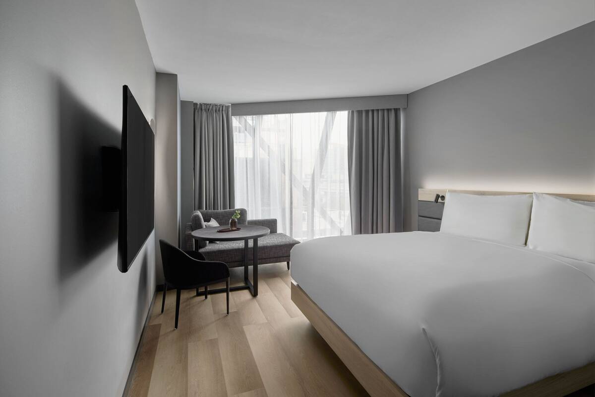 A modern hotel room with a large bed, a desk with a chair, a flat-screen TV, and a window with curtains.
