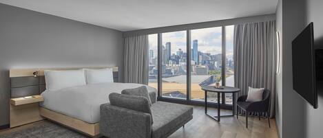 A modern hotel room with a large bed, a seating area, a small table, and a city view.