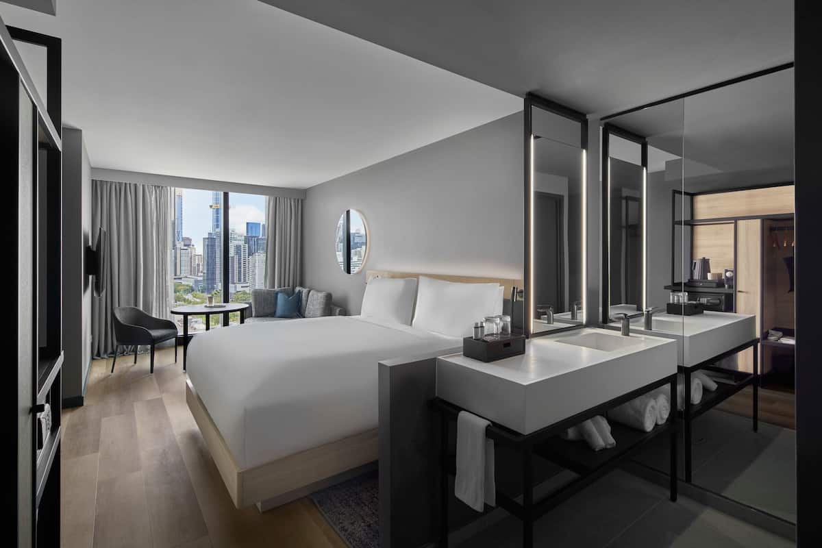 A modern hotel room with a large bed, a separate bathroom with a vanity, and a city view through large windows.