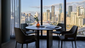 Una habitación moderna con un gran ventanal que ofrece vista panorámica de la ciudad, una mesa redonda con sillas y un florero con planta.
