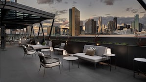 Una terraza en la azotea con mobiliario moderno, vistas del horizonte urbano y una estructura de vidrio.