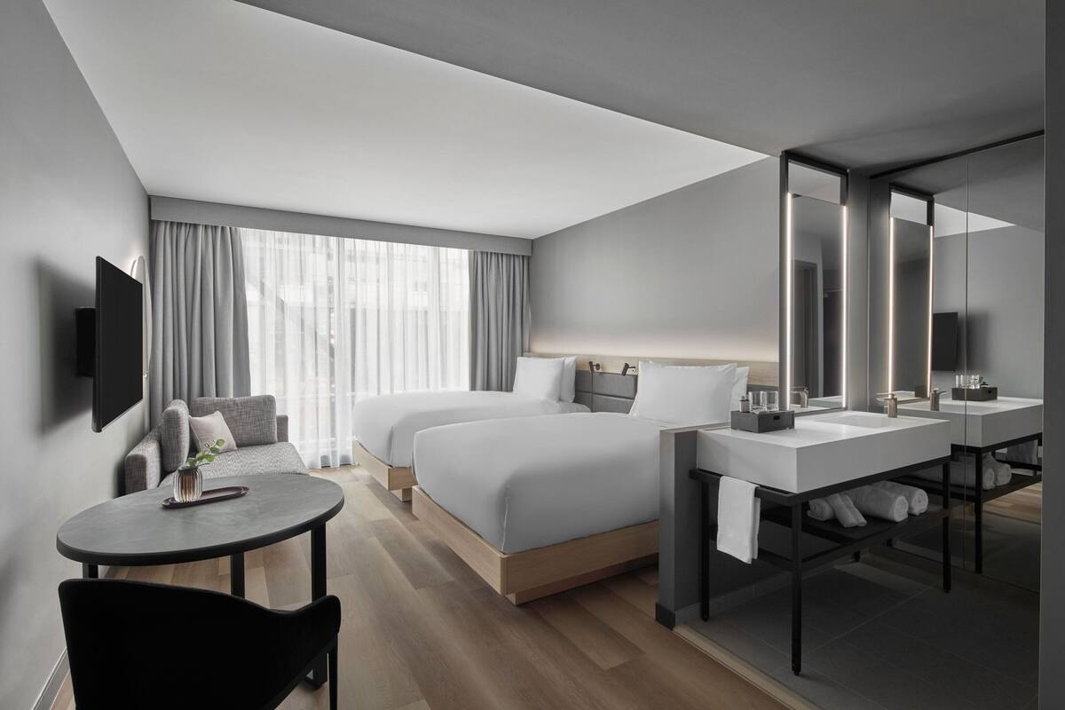 Habitació d'hotel moderna amb un llit gran, dues taules de nit, escriptori i una cadira. Hi ha una televisió penjada a la paret i una finestra gran amb cortines.
