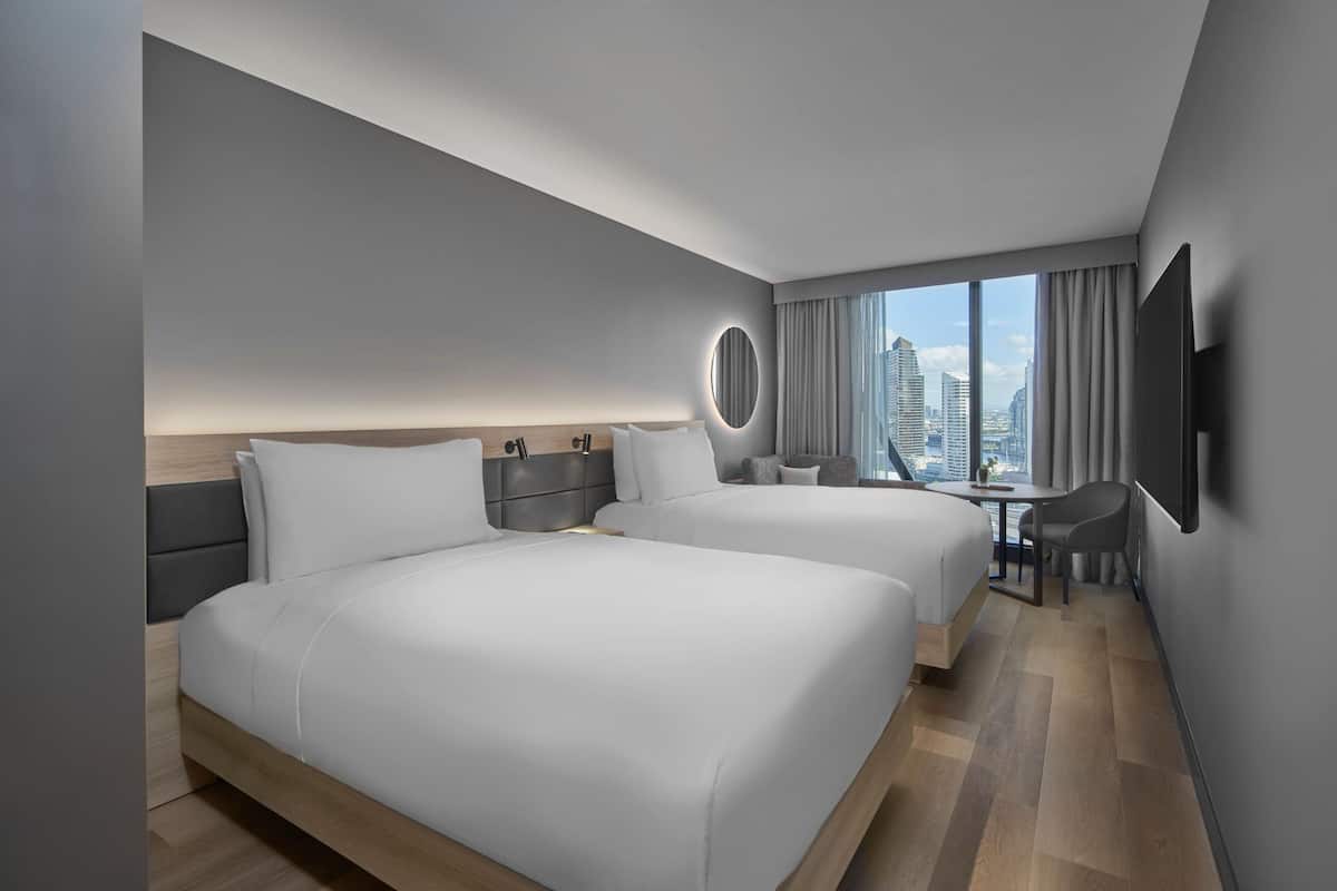 A modern hotel room with two beds, a desk, and a large window offering a city view.