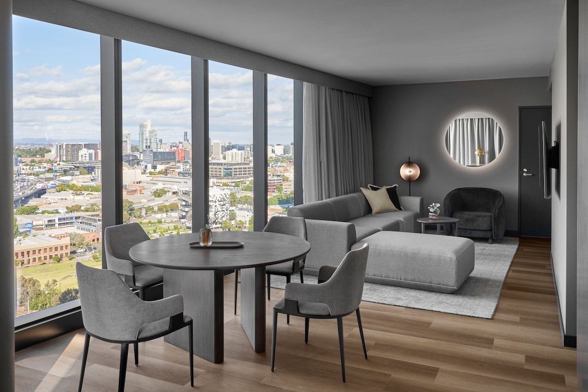 A modern living room with a large window, a round mirror, a sofa, a coffee table, and a dining area with chairs.