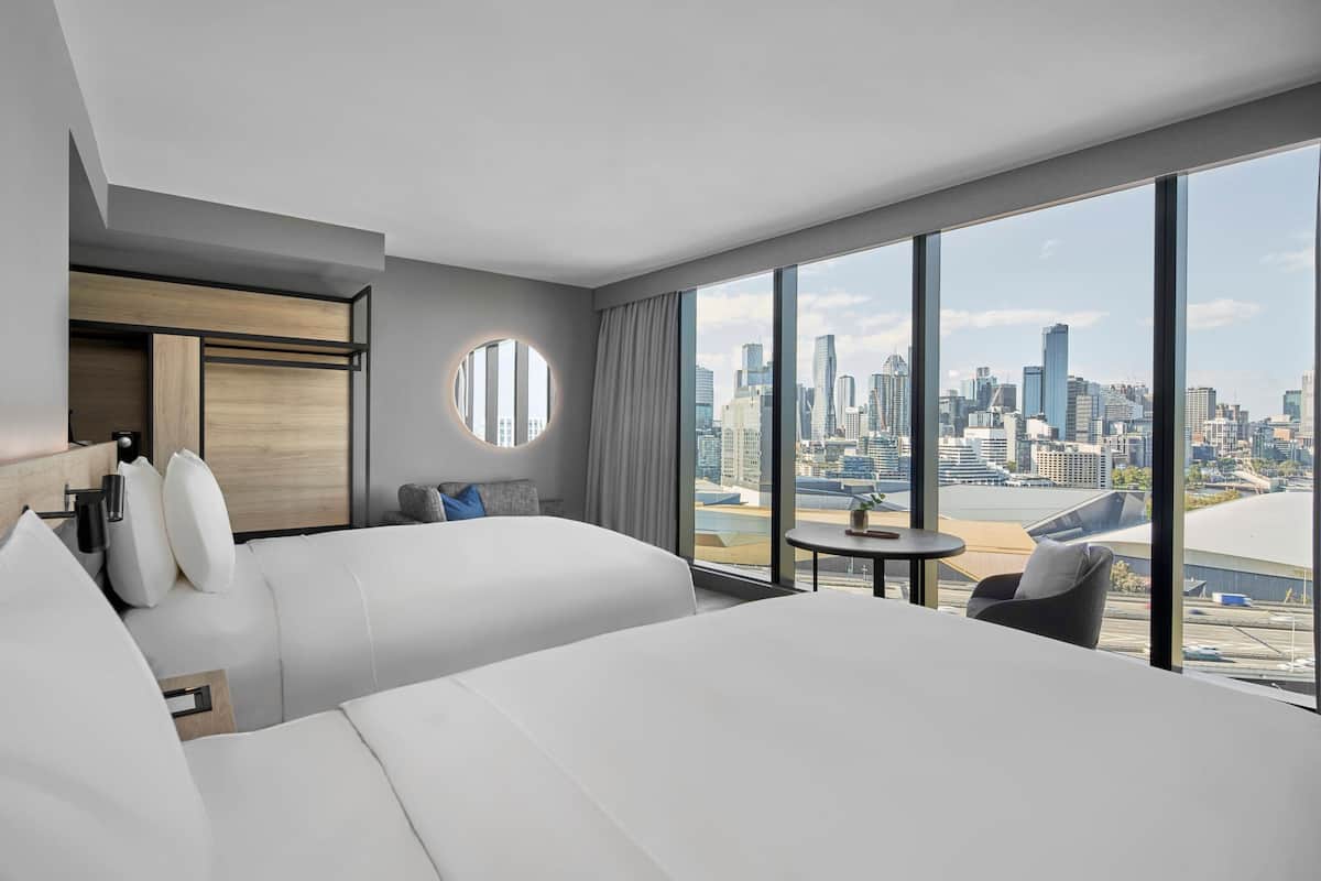 A modern hotel room with a large bed, a round mirror, a small table, and a view of a city skyline.