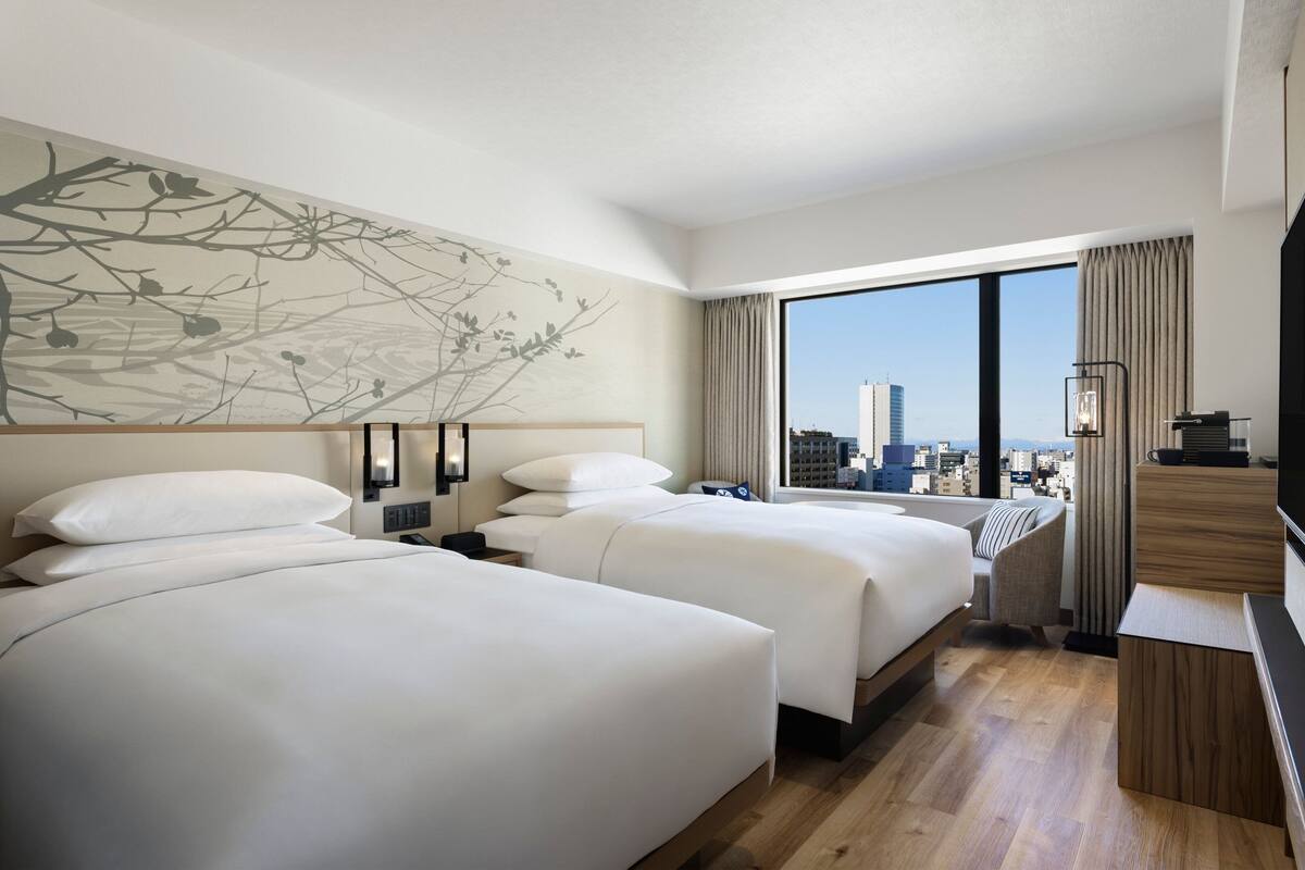 A hotel room with two beds, a large window with a city view, a wall art piece, and a TV on a wooden stand.