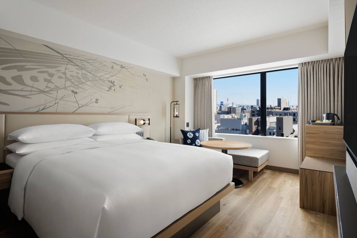 A modern hotel room with a large bed, a small table, a TV, and a city view.