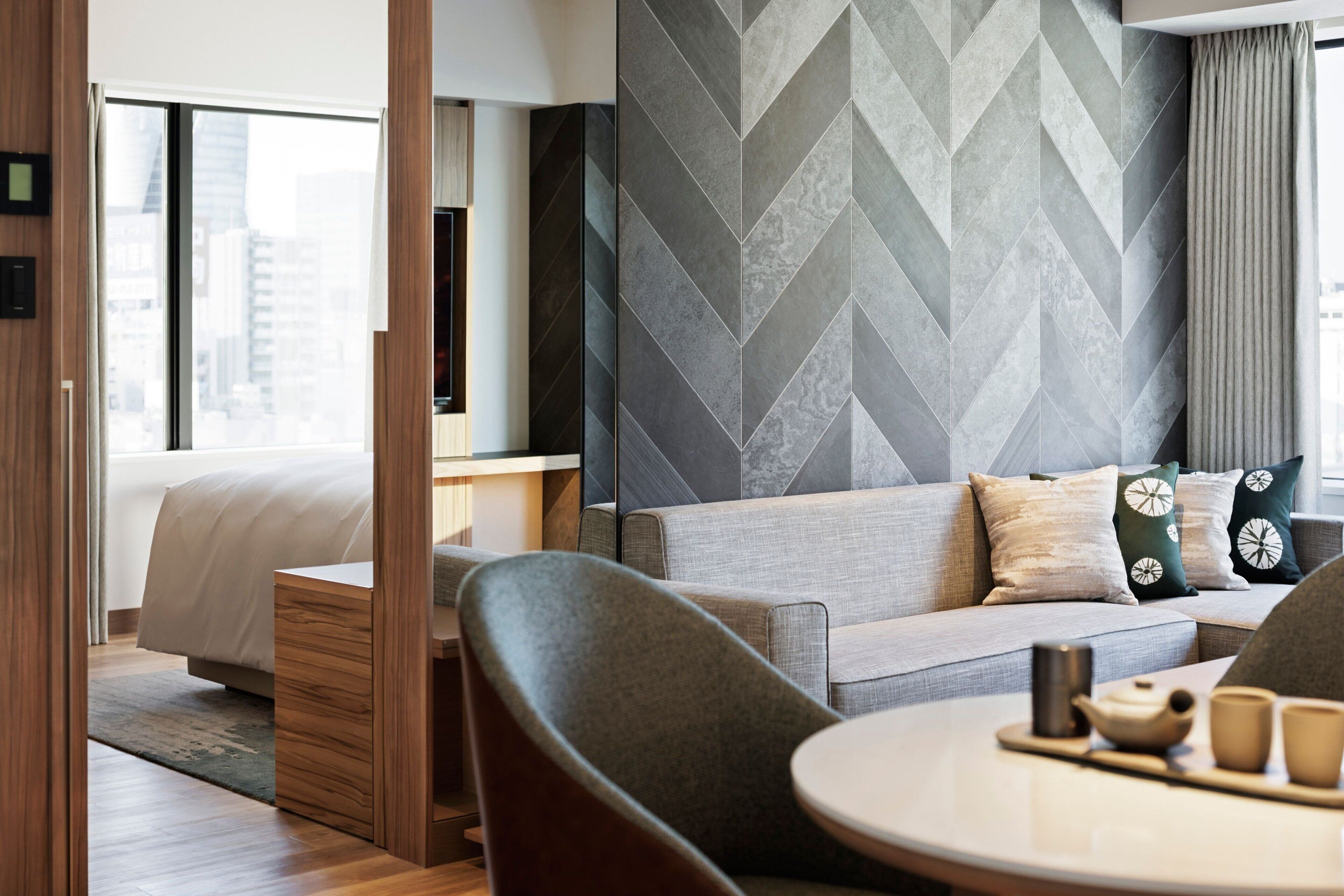 Habitación de hotel moderna con sofá, una mesa de comedor con tetera y tazas, y vista a la ciudad a través de grandes ventanales.