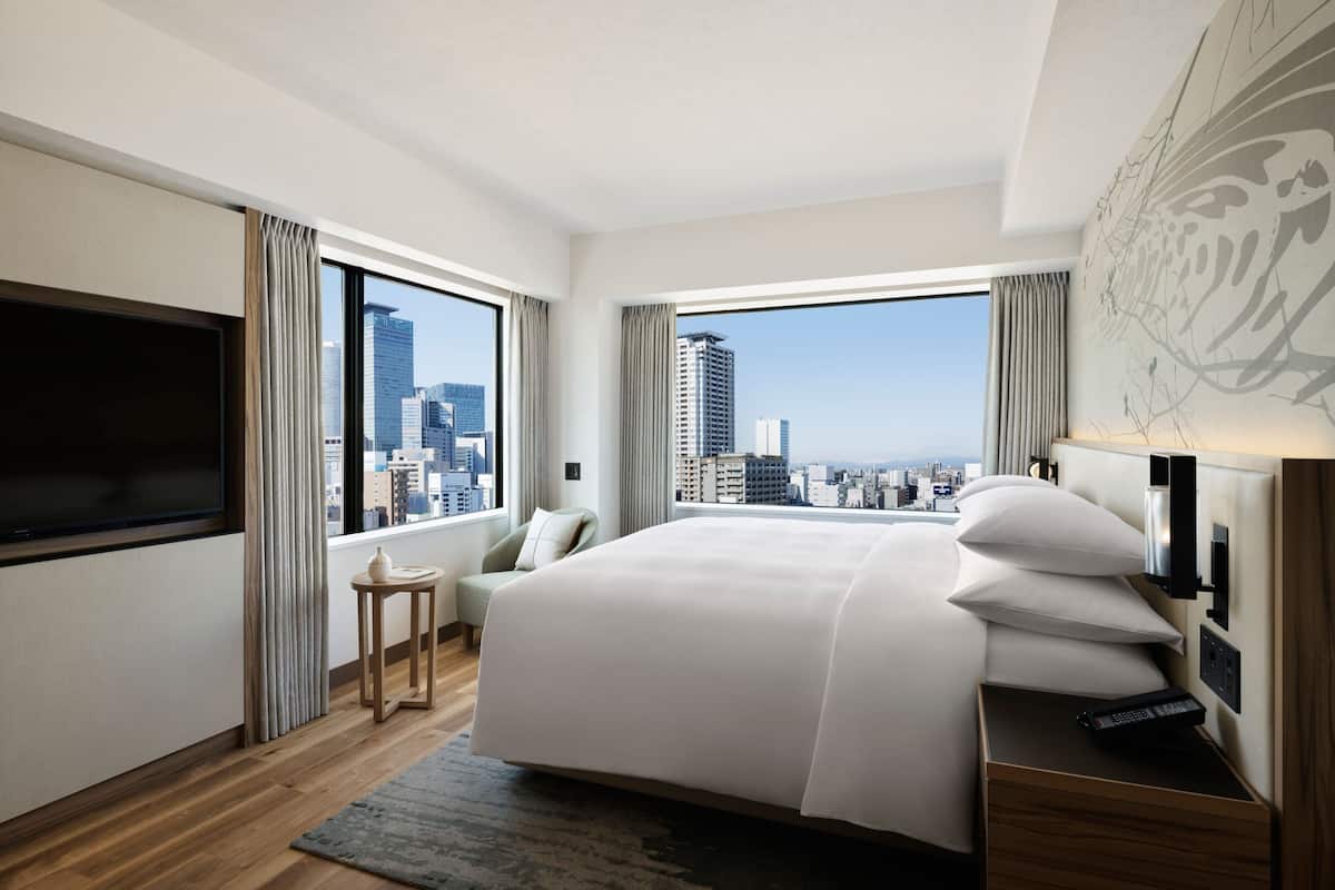 A modern hotel room with a large bed, a TV, a bedside table, and a view of the cityscape.