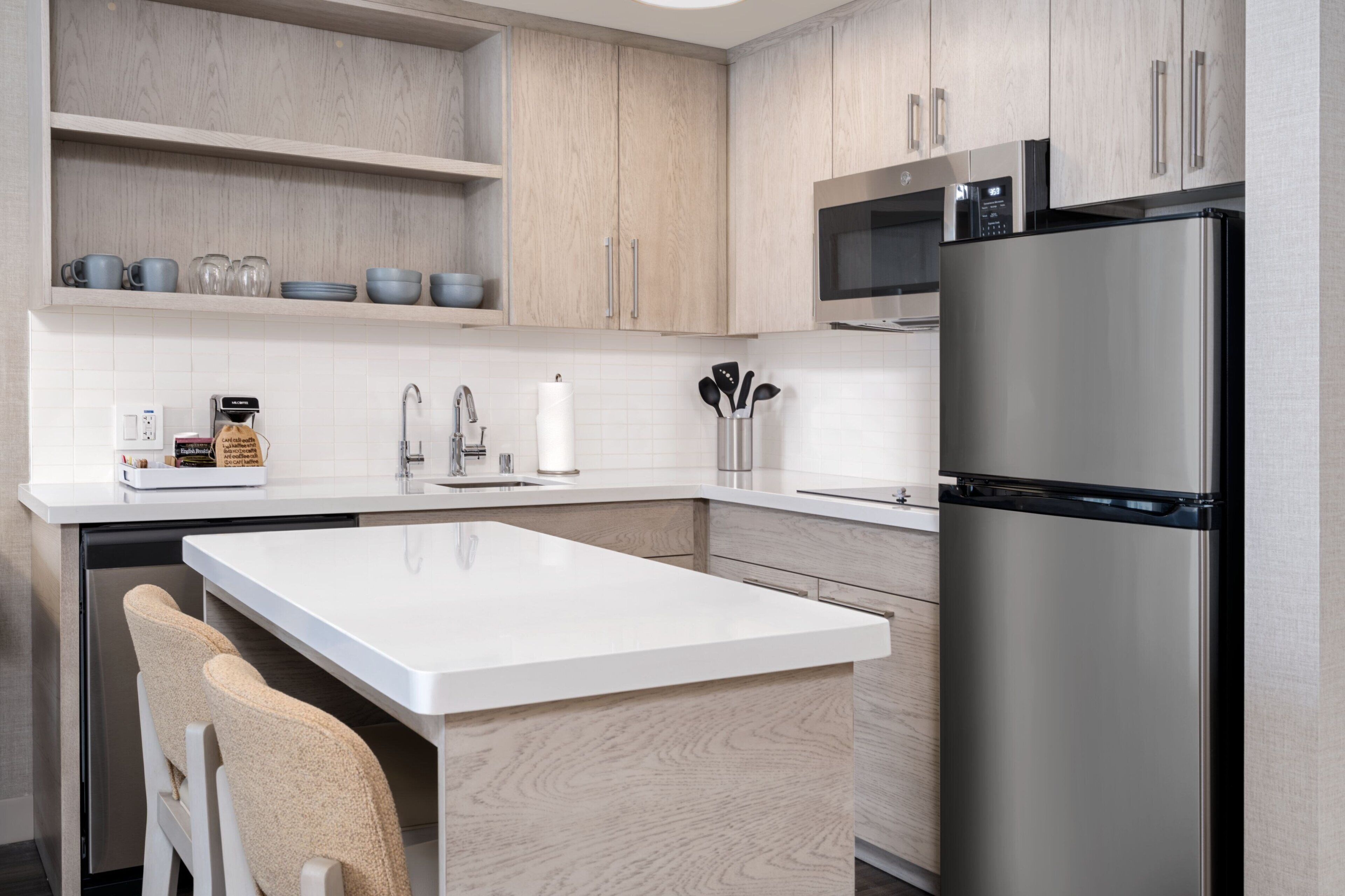 A modern kitchen with light wood cabinetry, a stainless steel refrigerator, a microwave, and a white countertop.