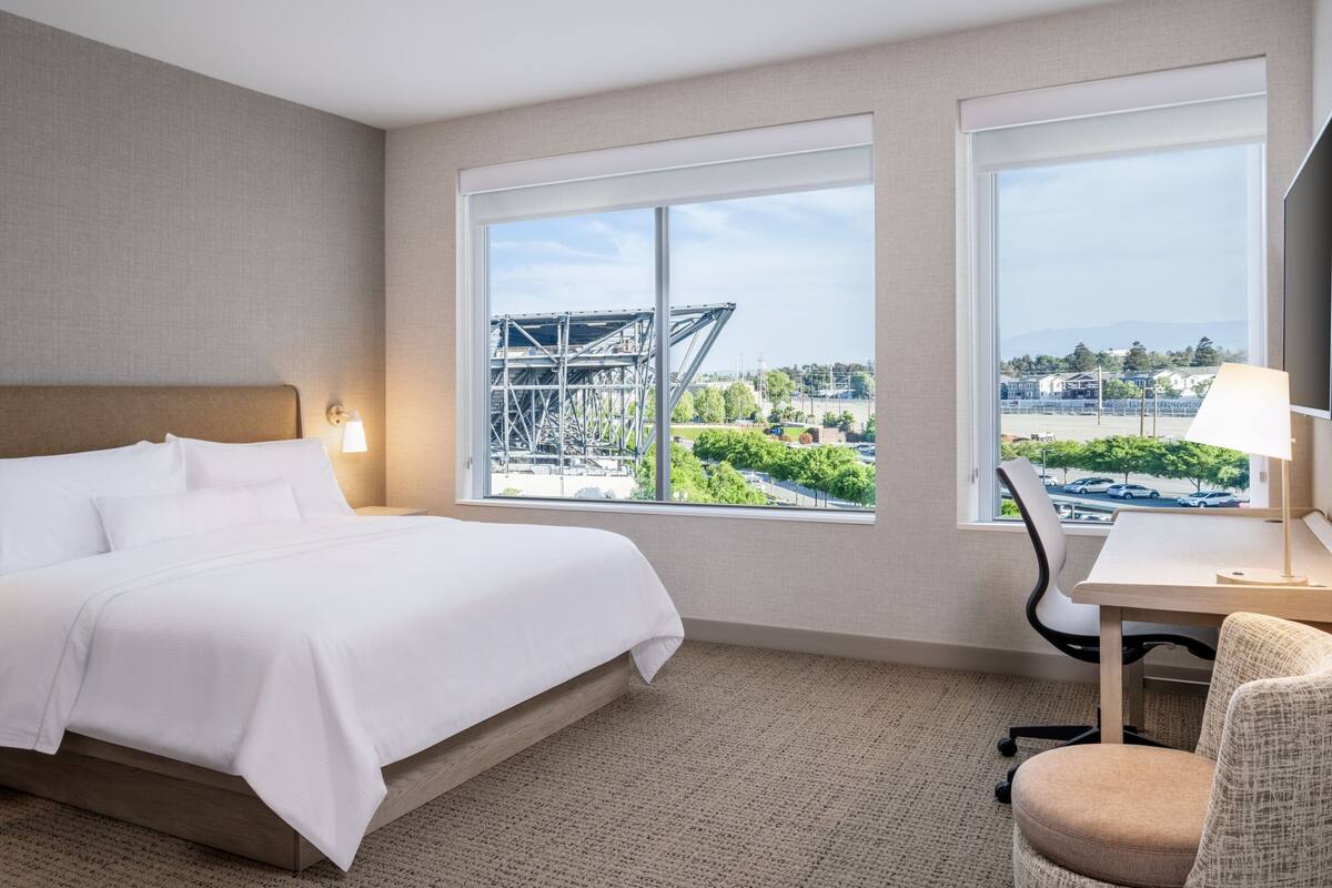 A hotel room with a bed, a desk, and two windows offering a view of a construction site and greenery.