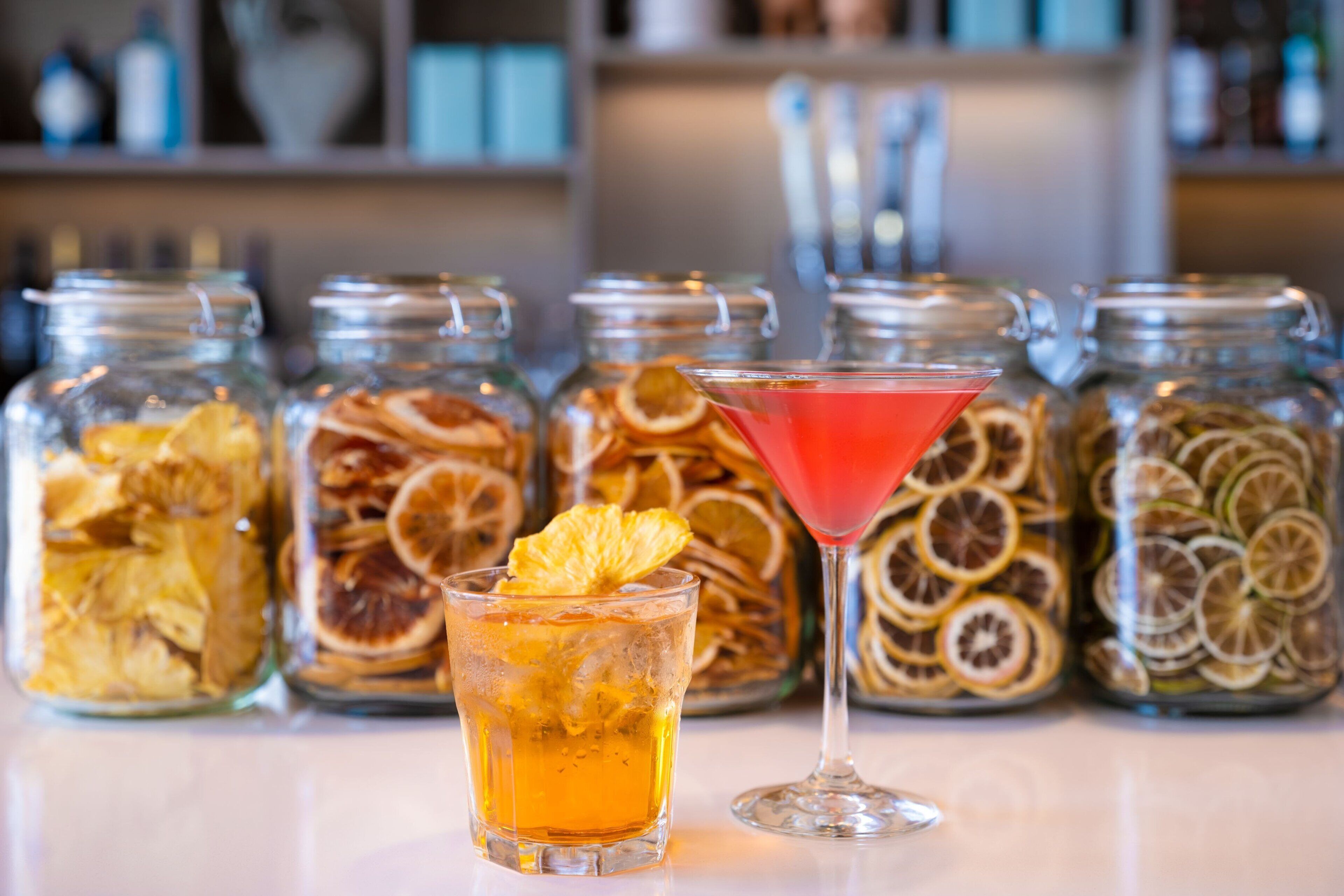 A bar area with a martini glass and a cocktail in a glass, surrounded by jars of dried fruit slices.