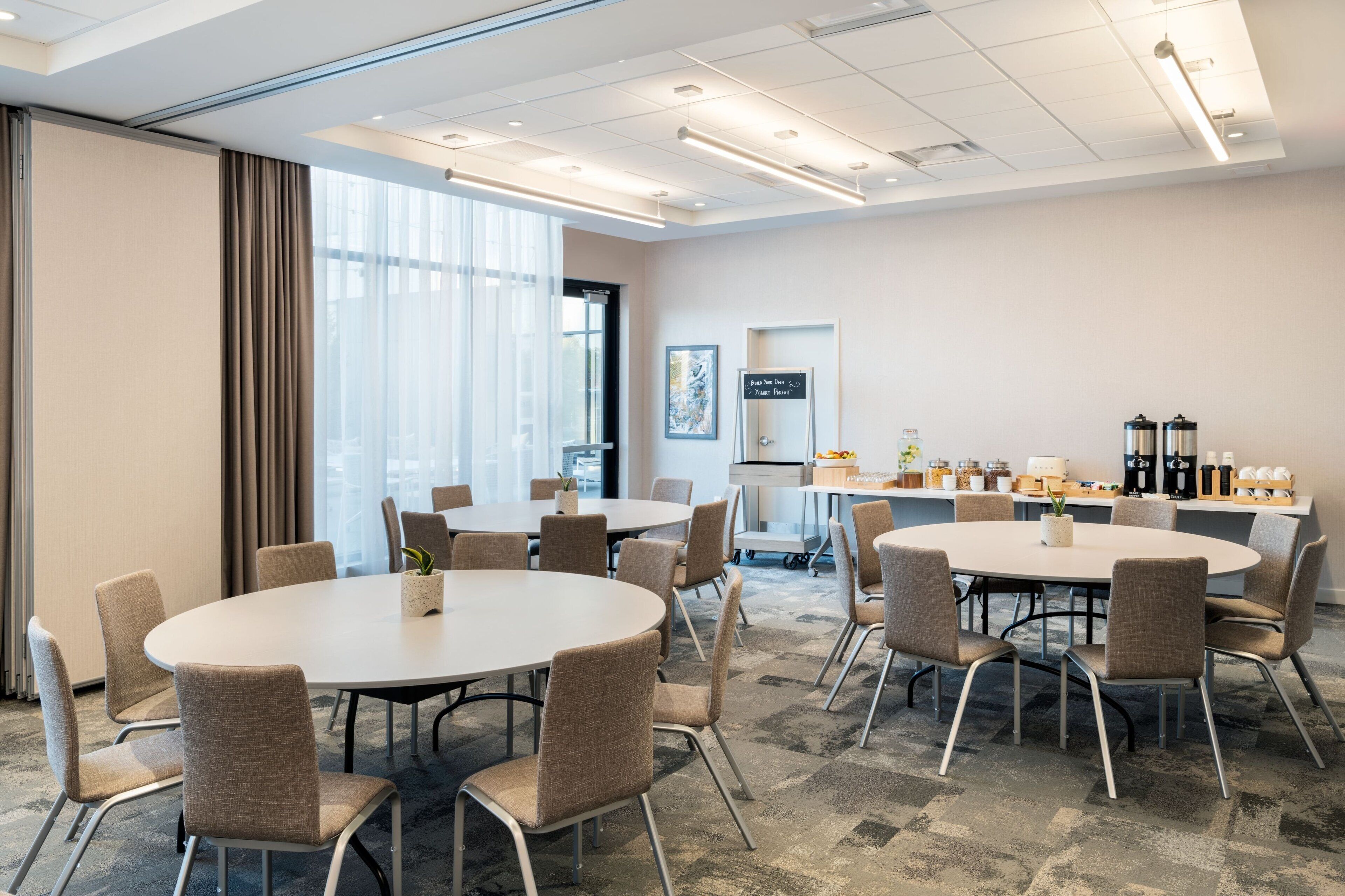 A conference room with a round table, multiple chairs, a coffee station, and a large window with curtains.
