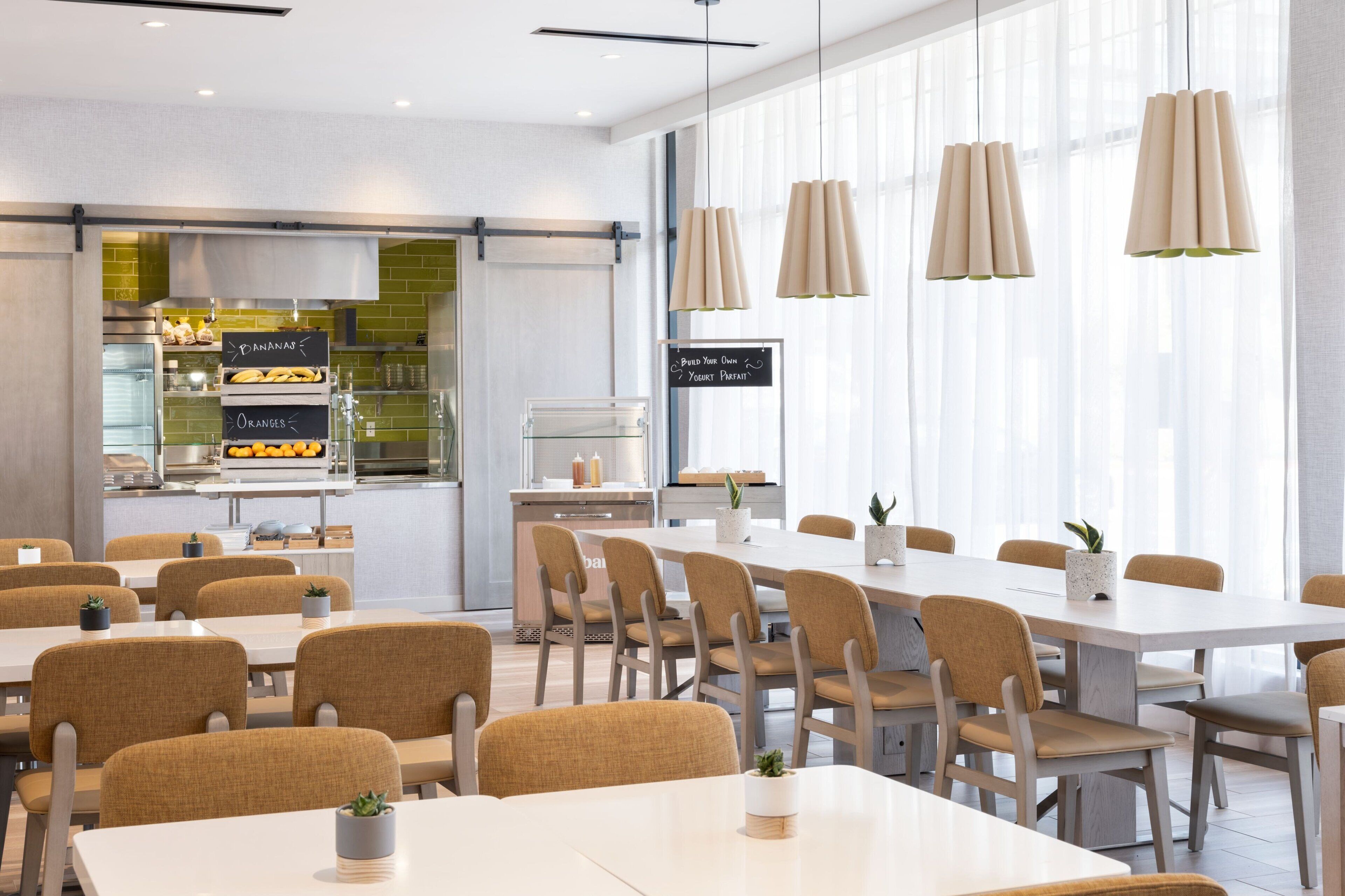 Un espace de salle à manger moderne avec des tables et des chaises blanches, une cuisine en arrière-plan et des luminaires suspendus.