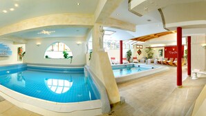 An indoor swimming pool with a clear view of the pool area, a separate smaller pool, and a lounge area with red pillars and seating. - Votters Hotel (Kaprun)