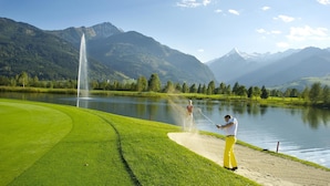 A golf course with a fountain, a lake, and mountains in the background. - Votters Hotel (Kaprun)