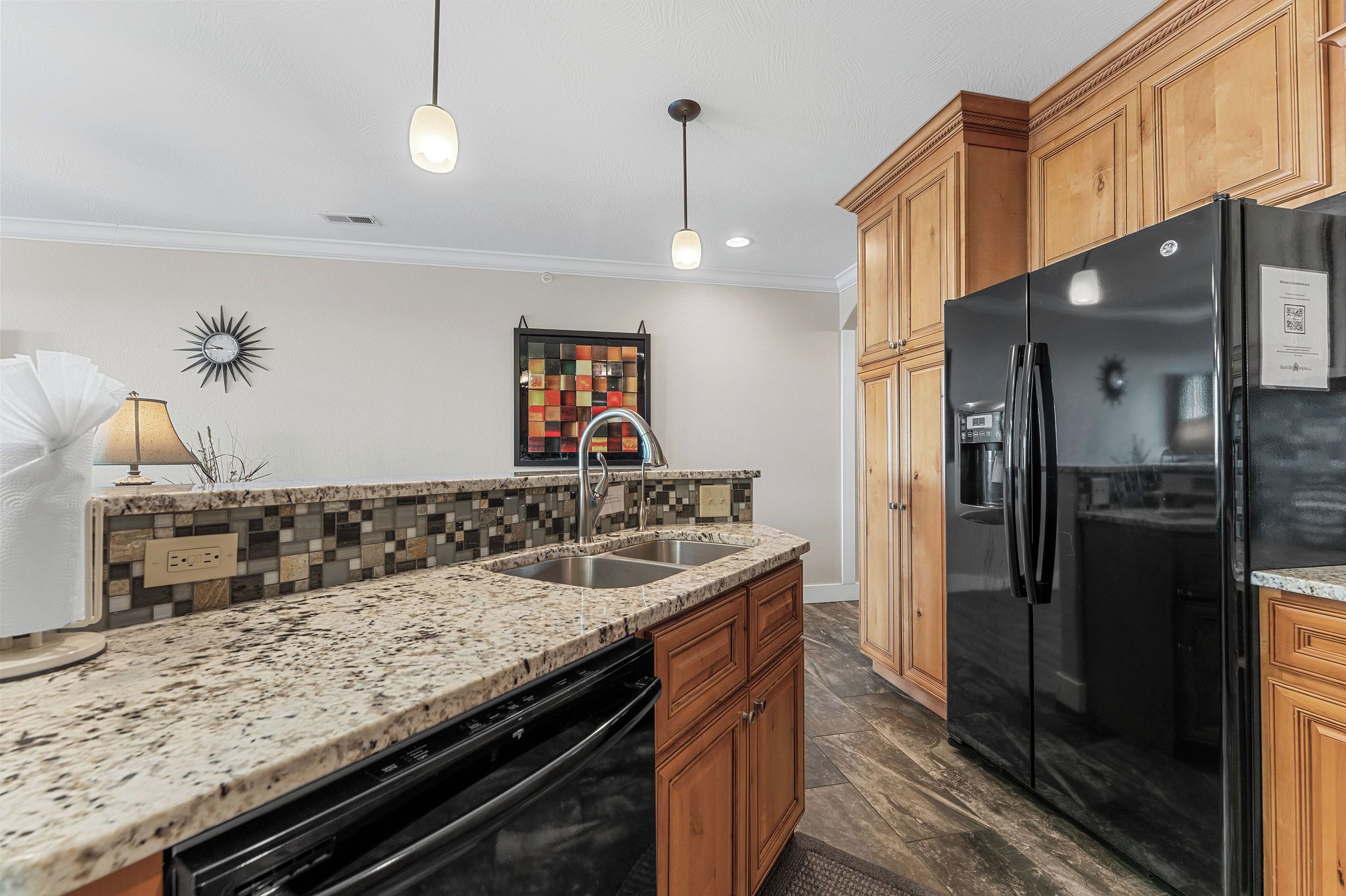 Una cocina con encimeras de granito, armarios de madera, refrigerador negro e iluminación colgante.