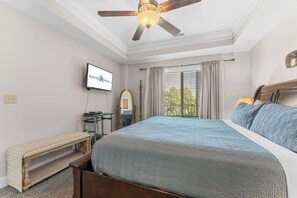 Ein Schlafzimmer mit einem Bett, einem Deckenventilator, einem Fernseher, einem Fenster mit Vorhängen und einer Sitzbank.