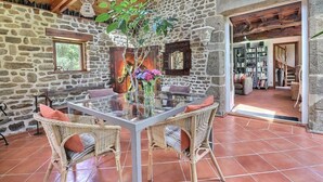 Una habitación de paredes de piedra con una mesa de vidrio y sillas de mimbre, un jarrón con flores y una estantería al fondo.