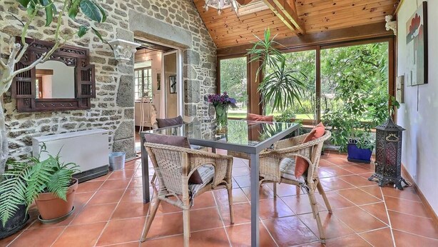 Un interior rústico de piedra con una mesa de vidrio y sillas de mimbre, rodeado de plantas en macetas y vistas al verdor exterior.