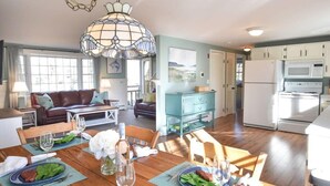 A spacious living area with a dining table set for two, a sofa, and a kitchen with white appliances. - Peace and Quiet & Spacious Back Deck (Wellfleet)