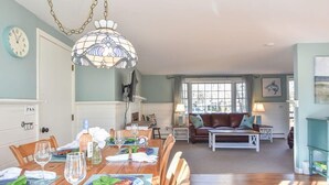 A dining area with a wooden table set for a meal, a chandelier, and a view of a living room with a sofa and a lamp. - Peace and Quiet & Spacious Back Deck (Wellfleet)