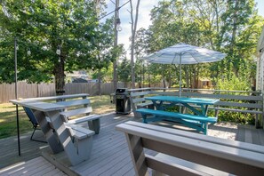 A deck with picnic tables and an umbrella, surrounded by trees and a wooden fence. - Peace and Quiet & Spacious Back Deck (Wellfleet)