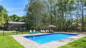 A rectangular swimming pool with a brick border, surrounded by a black fence, and a grassy area with lounge chairs and a red umbrella. - Heated In-Ground Pool and New Central AC! (Brewster)