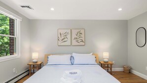 A neatly made bed with a folded towel and a pillow, two wall-mounted lamps, a window with a view of trees, and a ceiling vent.