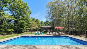 A rectangular swimming pool with a tiled edge, surrounded by a brick patio. There are lounge chairs and a red umbrella on the right side. - Heated In-Ground Pool and New Central AC! (Brewster)