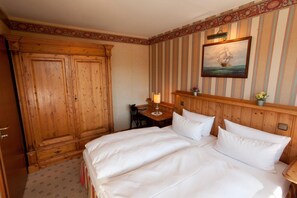 Habitación de hotel con un gran armario de madera, cama con ropa blanca, escritorio con lámpara y un cuadro colgado en la pared que muestra un barco de vela.
