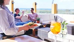 A person holding a plate of food at a table with a drink in a glass, with a view of the ocean in the background. - Moby Dick Wustrow (Ostseebad Wustrow)