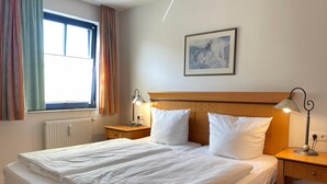 A hotel room with a double bed, two bedside tables with lamps, a window with curtains, a radiator, and a painting on the wall. - Moby Dick Wustrow (Ostseebad Wustrow)