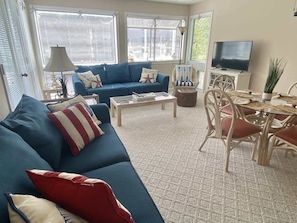A living room with a blue sofa, a coffee table, a dining table with chairs, a TV, and a lamp. - Lovely 2BR,Pool,M28 (Myrtle Beach)