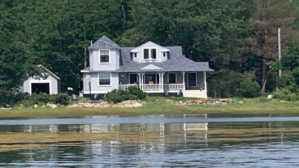 Une maison à deux étages avec un porche, entourée d’arbres et bordée par un plan d’eau.