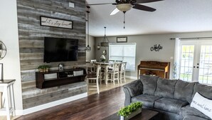 Un salon doté d’un parquet en bois foncé, d’un canapé gris, d’une table basse agrémentée d’une plante, d’un téléviseur fixé à un mur en bois, d’un coin repas avec une table et des chaises, et d’un piano.
