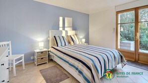 Quarto com cama, mesa de cabeceira, abajur e vista do exterior por uma porta.