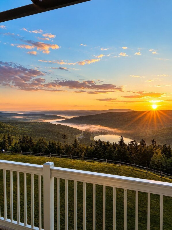 A scenic view from a balcony overlooking a valley with a sunrise, a lake, and a forested area.