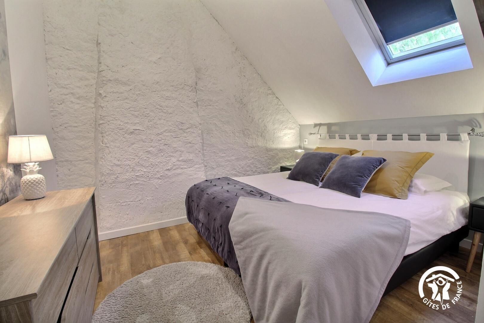 A bedroom with a sloped ceiling, a skylight, a bed with pillows, a nightstand with a lamp, and a wooden dresser.