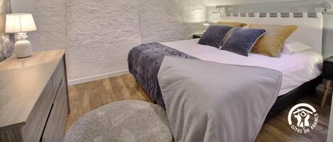 A bedroom with a sloped ceiling, a skylight, a bed with pillows, a nightstand with a lamp, and a wooden dresser.