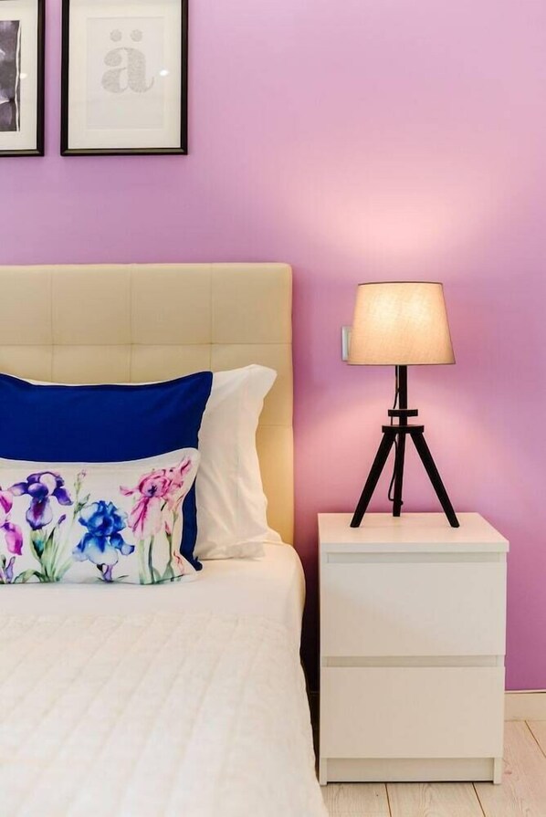 Una cama con un cojín floral, una lámpara de noche en una mesita blanca y fotografías enmarcadas en la pared.