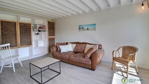 Een woonkamer met een bruine leren bank, een witte stoel, een rieten stoel, een glazen salontafel en een schilderij aan de muur.