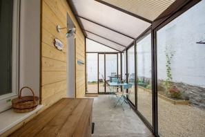 Un porche cubierto con una mesa y sillas de madera, una cesta de mimbre y un ventanal que da a un jardín.