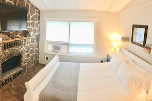 A bedroom with a stone fireplace, a bed with white linens, a window with blinds, and a wall-mounted TV. - Bright & Clean Chalet with Hot Tub (Acme)