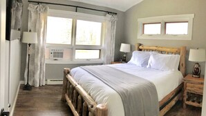 A bedroom with a wooden bed frame, a large window with curtains, a bedside table with a lamp, and a wall-mounted heater. - Bright & Clean Chalet with Hot Tub (Acme)
