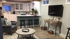 Un salon moderne avec un canapé gris, une table basse ronde et un téléviseur fixé au mur. On y trouve également un coin cuisine avec des meubles blancs et un petit îlot.