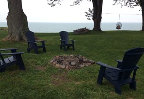 A grassy outdoor area with a fire pit, several Adirondack chairs, and a hanging swing chair.