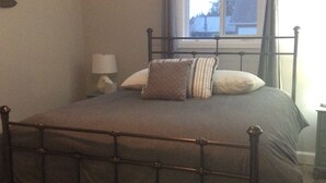 Une chambre à coucher avec un lit en métal, une fenêtre avec des rideaux, une lampe de chevet et une vue sur une maison à l’extérieur.